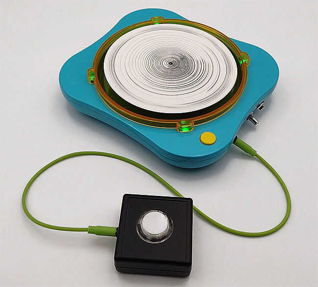 Turquoise spin art machine with four green lights around it, and a spinning white disc of paper with black concentric part drawn circles. In front an accessibility switch.