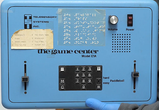 The Game Center by Telesensory Systems Inc. (TSI) from 1978. A light blue aluminium box with number pad, two vertical sliders either side of the box and various other controls. Braille and printed game list from 0-7.