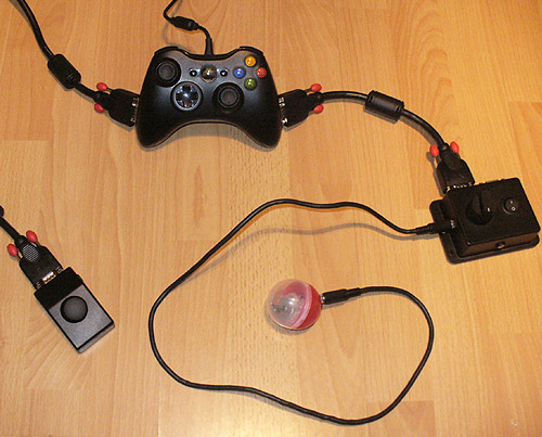Image of a black adapted Xbox 360 joypad, connected via chunky Lindy SVGA cables to a box with a dial, on/off switch and two sockets. One socket for a switch, the other for a Vibro Capsule (Red and clear one with motor inside attached). On the other side an external thumbstick in a small sloping black case is attached.